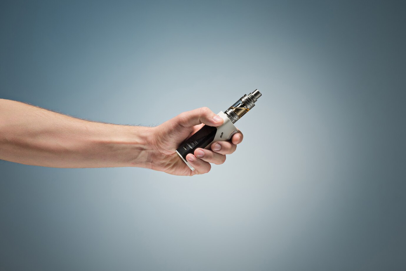 A person’s hand is holding a silver vape device against a gradient blue background—an image that captures the sleek appeal essential for scaling a vape business. The cylindrical vape features a clear tank and mouthpiece on top.